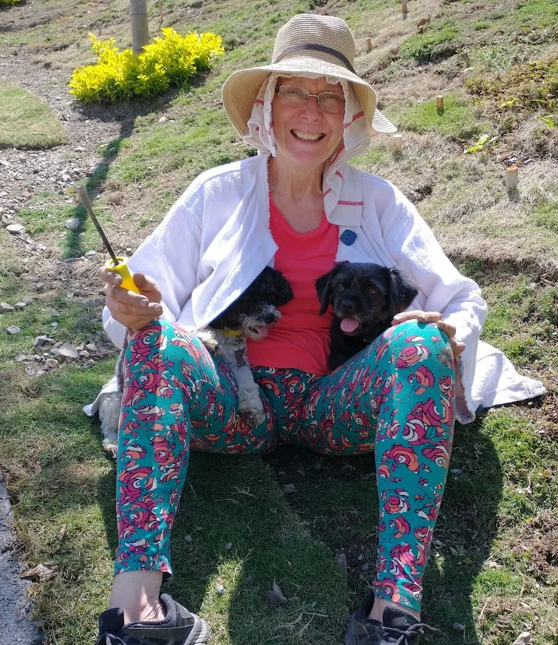 A smiling woman in a sun hat sits on the grass with her two small dogs.