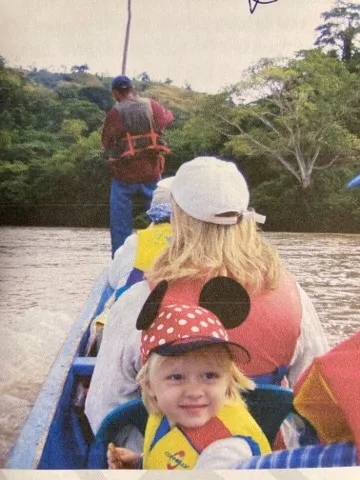 A young child in a life jacket smiles from a small river boat, with family seated in front.