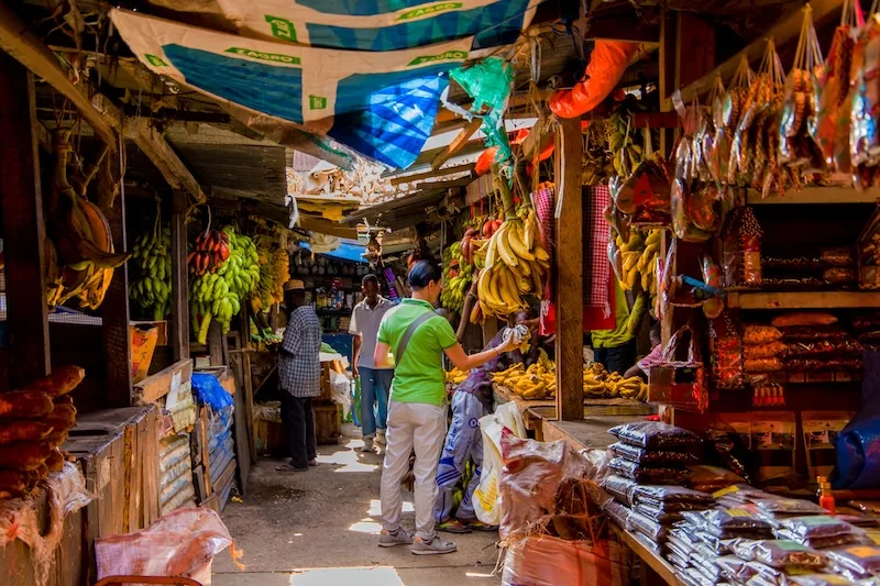 Shoppers walk down a covered market lane lined with bananas and dried goods.