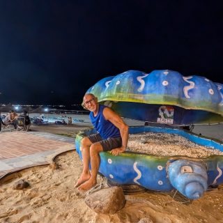 A smiling man sits on a giant painted seashell sculpture on the beachfront in Da Nang at night.