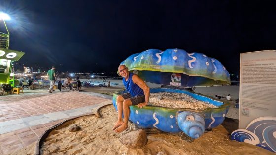 A smiling man sits on a giant painted seashell sculpture on the beachfront in Da Nang at night.