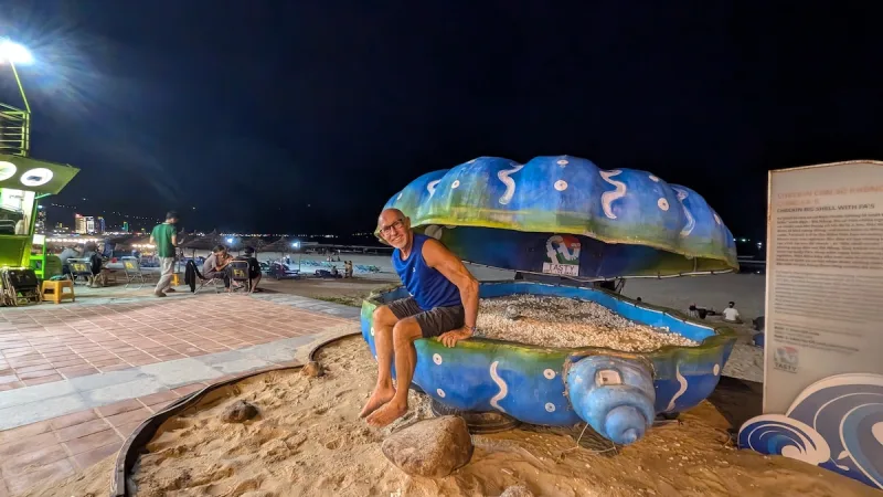 A smiling man sits on a giant painted seashell sculpture on the beachfront in Da Nang at night.