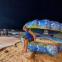 A smiling man sits on a giant painted seashell sculpture on the beachfront in Da Nang at night.