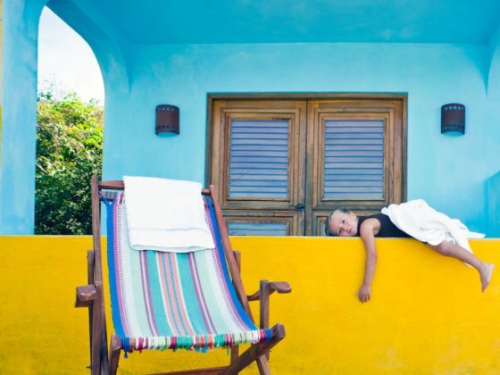 A young girl rests on a bright yellow balcony beside a striped deck chair.