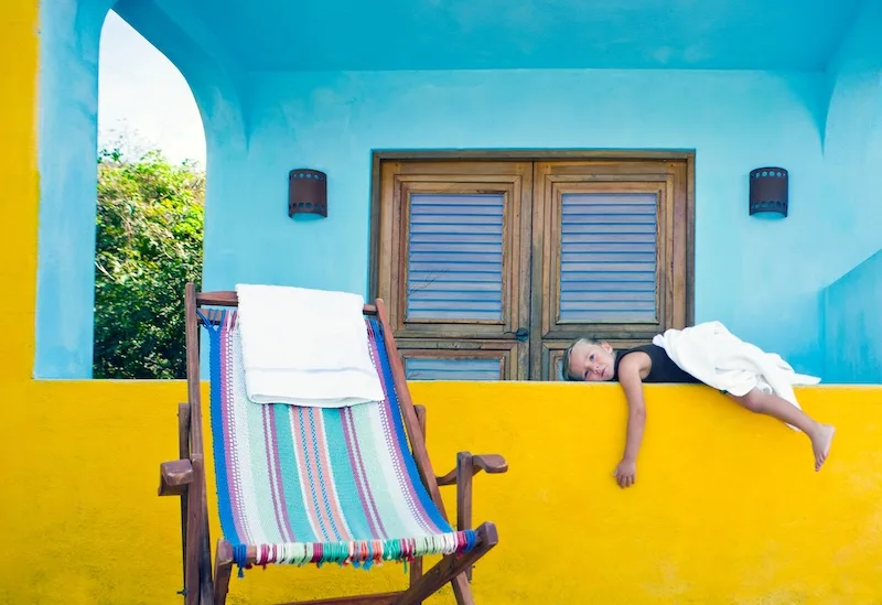 A young girl rests on a bright yellow balcony beside a striped deck chair.