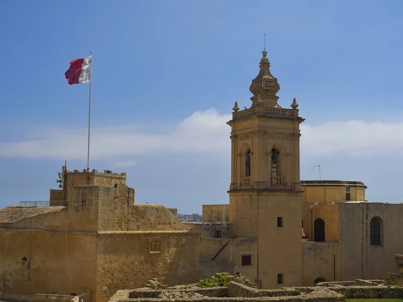 The Maltese flag flies over the stone walls and bell tower of the Citadel in Victoria, Gozo.