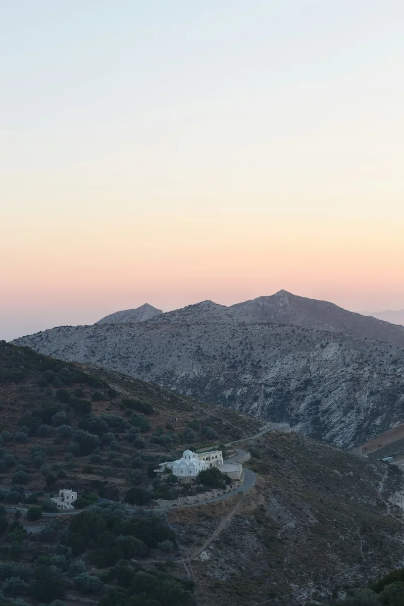 A white chapel on a winding mountain road on a Greek island at dusk.