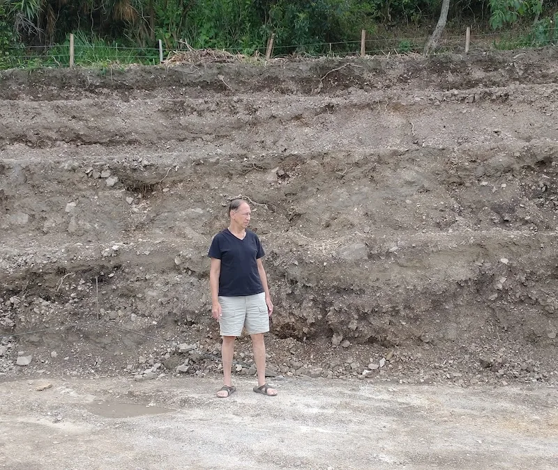 A man stands in front of the steep, rocky cut dug into the hillside to make a level building site.