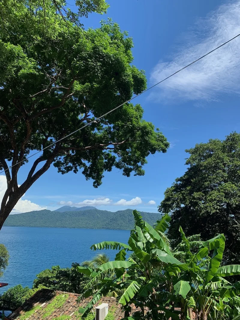 The blue water of Laguna de Apoyo framed by trees and banana plants, with volcanic hills behind, in Nicaragua.