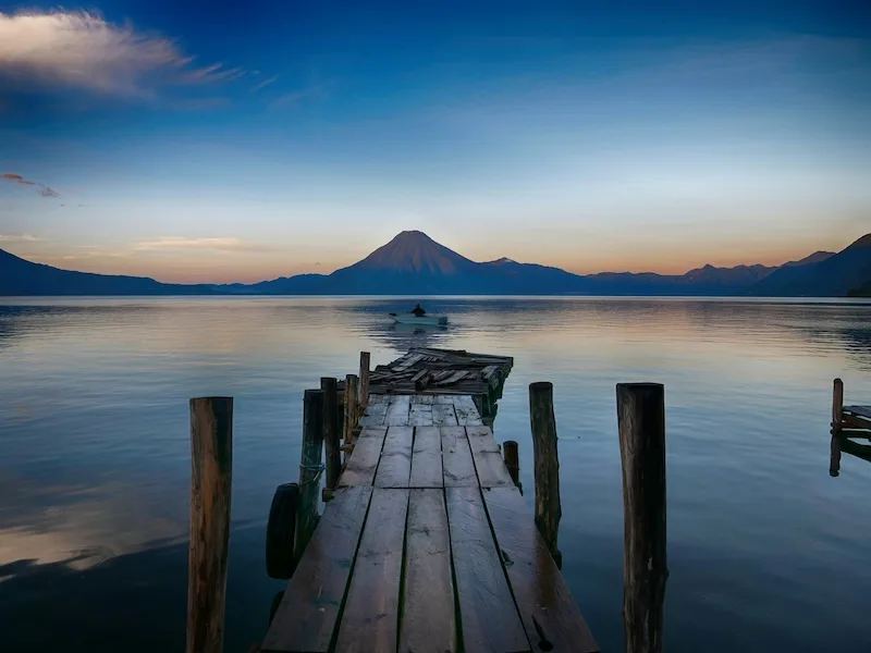 A weathered wooden dock reaches into still water at dawn, a volcano on the far shore: the view that sells living in Lake Atitlán