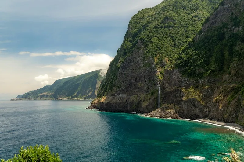 A waterfall drops from green sea cliffs into turquoise water on the north coast of Madeira.