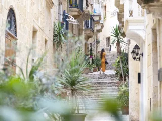 A woman in a yellow dress walks up a plant-lined limestone street in Malta, the country facing the first of the expat rule changes 2027 brings