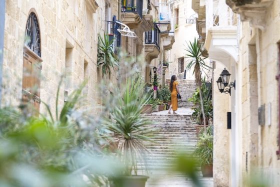 A woman in a yellow dress walks up a plant-lined limestone street in Malta, the country facing the first of the expat rule changes 2027 brings