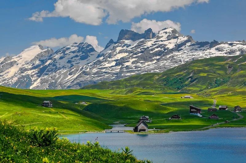 Chalets and a small chapel beside a mountain lake below snow-capped peaks in the Swiss Alps.