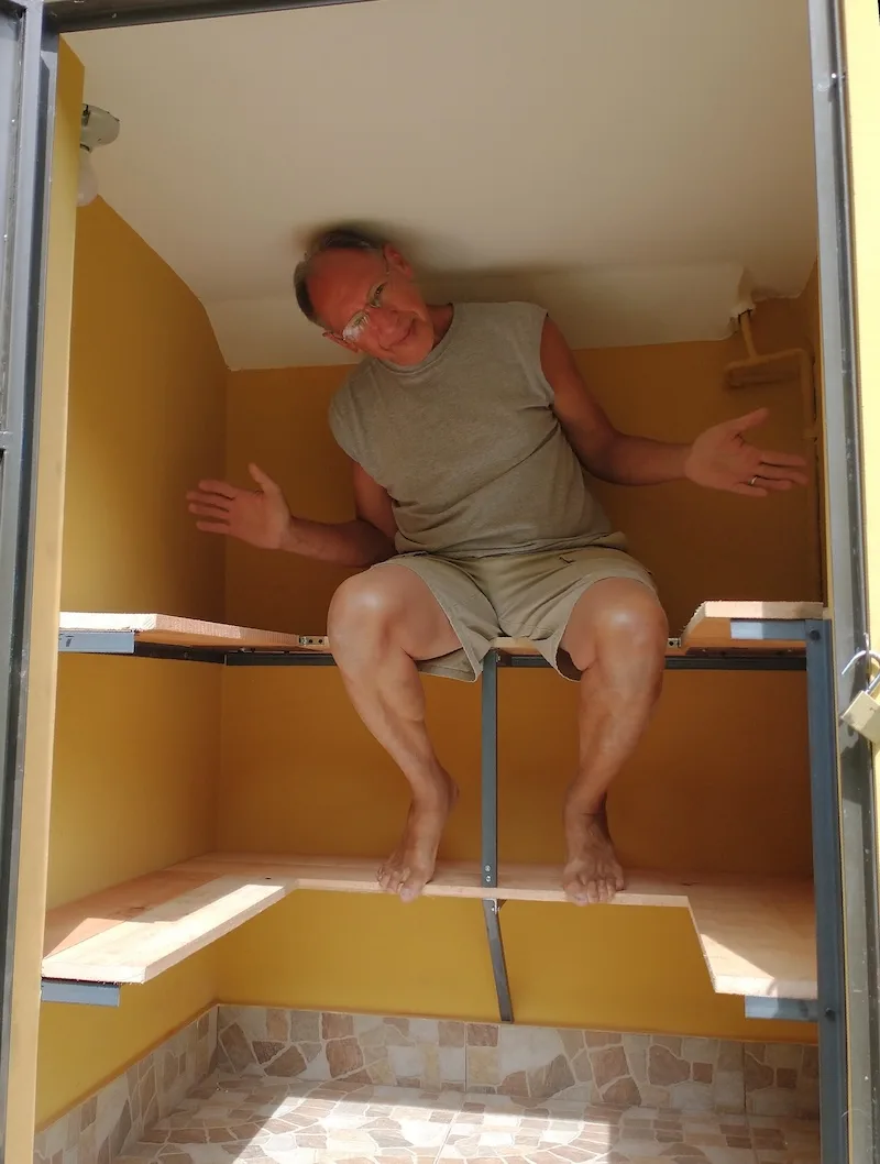 A smiling man sits on the new shelving inside a small yellow closet.