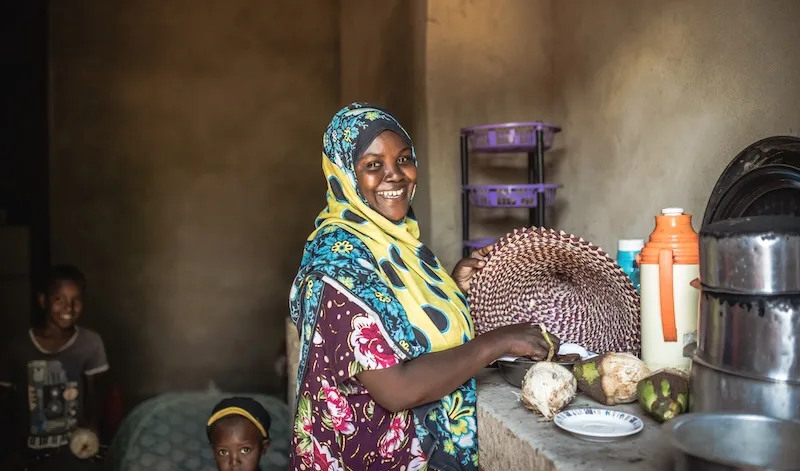 A smiling woman in a bright headscarf prepares food in her kitchen as two children look on.