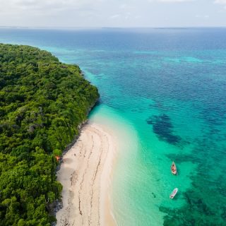 Aerial view of a white-sand beach and turquoise Indian Ocean water beside a forested stretch of Zanzibar coast, with two boats moored offshore