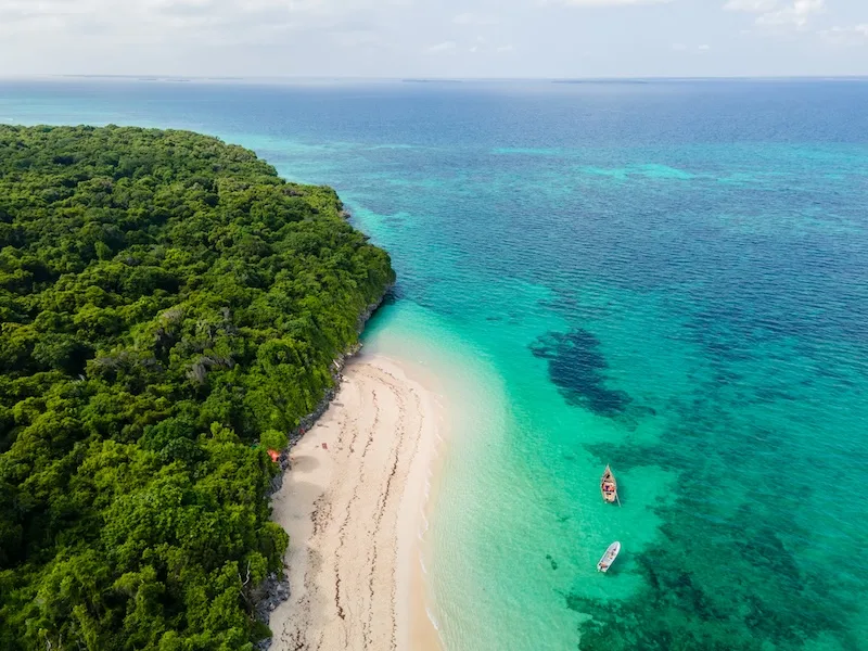 Aerial view of a white-sand beach and turquoise Indian Ocean water beside a forested stretch of Zanzibar coast, with two boats moored offshore