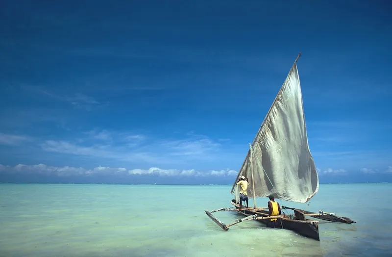 Two fishermen sail a traditional outrigger canoe across shallow turquoise water off Zanzibar.